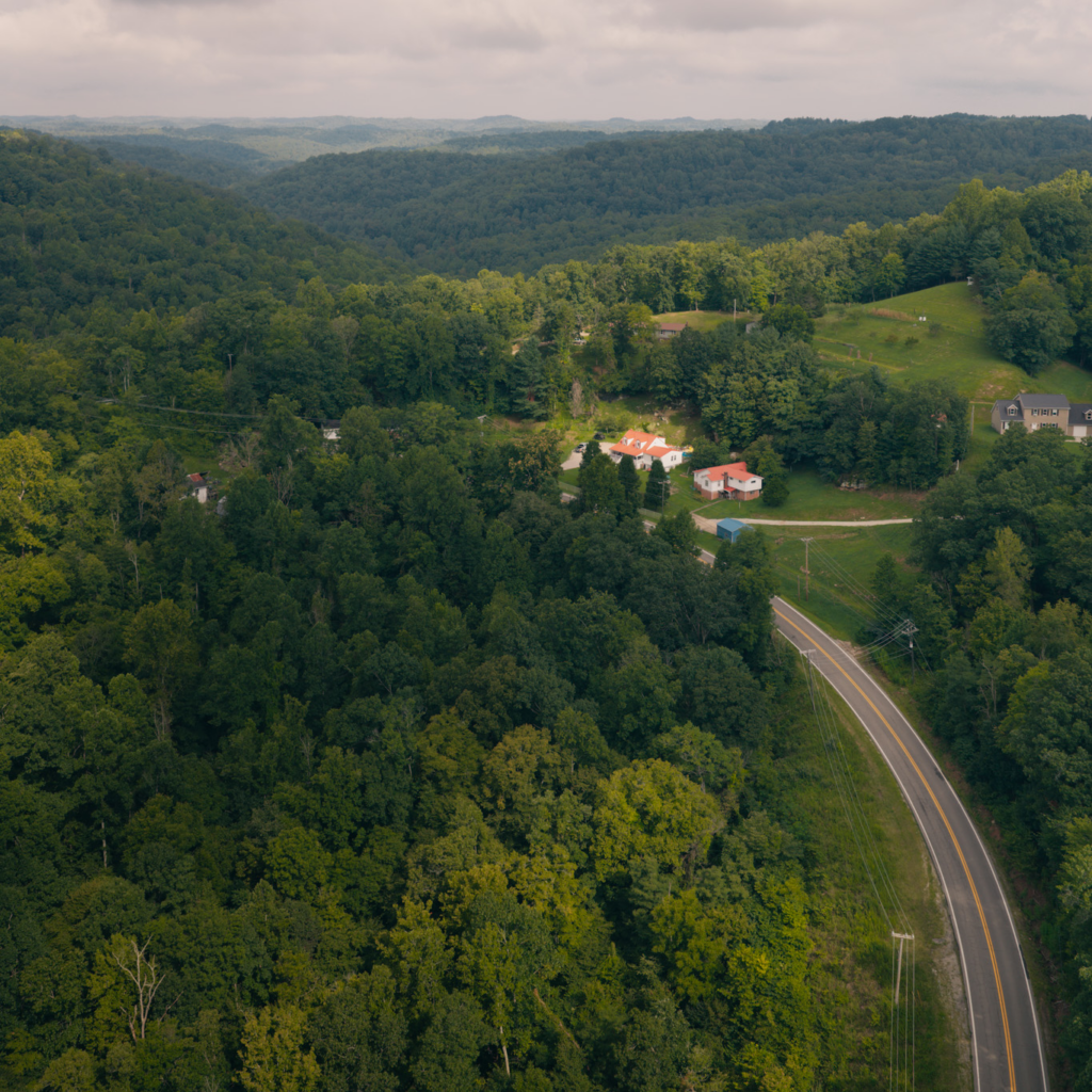 Drone image of a curving country road cutting through green trees. You can see a house with a red roof in the foreground with rolling hills behind it. 